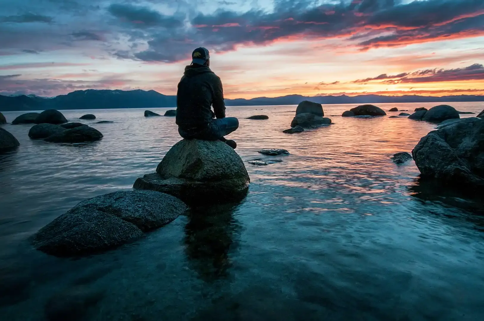Person meditating on a rock beside a lake at sunset